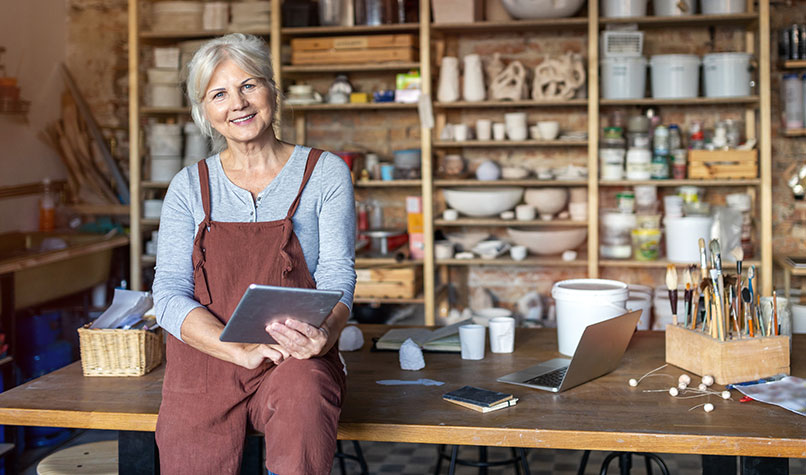 A craftswoman sat on a counter with an array of her products on shelves behind her.