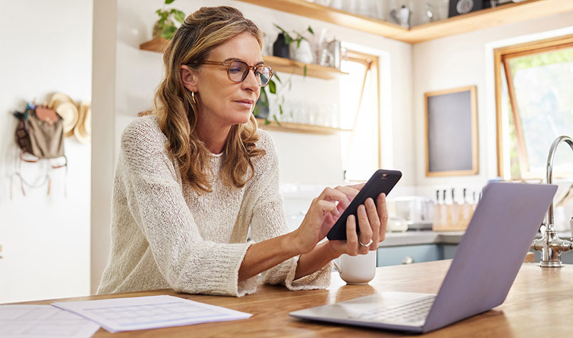 Woman looking at her phone while sat at a table with a laptop and documents