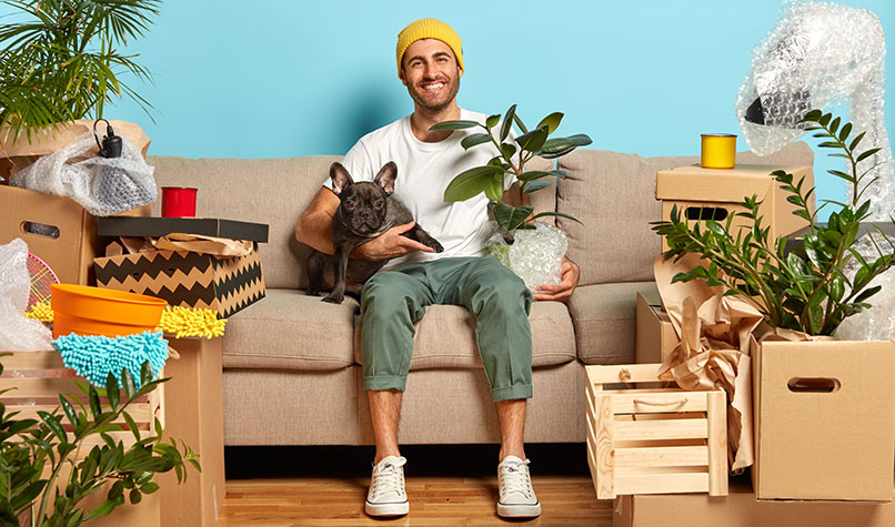 A man sat on a sofa holding a dog and surrounded by boxes