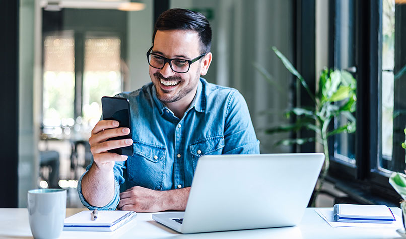 Man looking at his phone at a desk upon which he has his laptop open