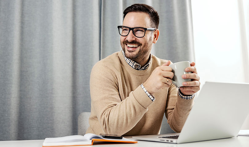 A man wearing spectacles and holding a cup sat at a desk