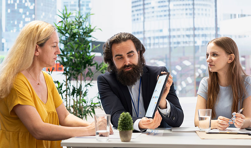 Bearded man showing a blonde woman a tablet computer while a dark haired woman looks one