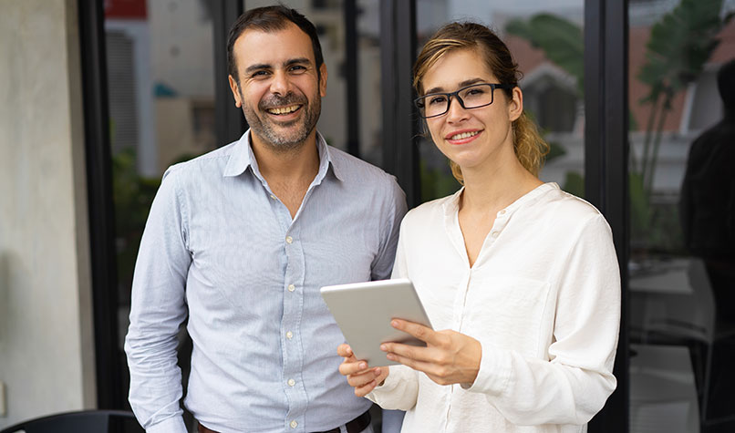A male and female in their 30s looking at the camera. The female is holding a small tablet computer.
