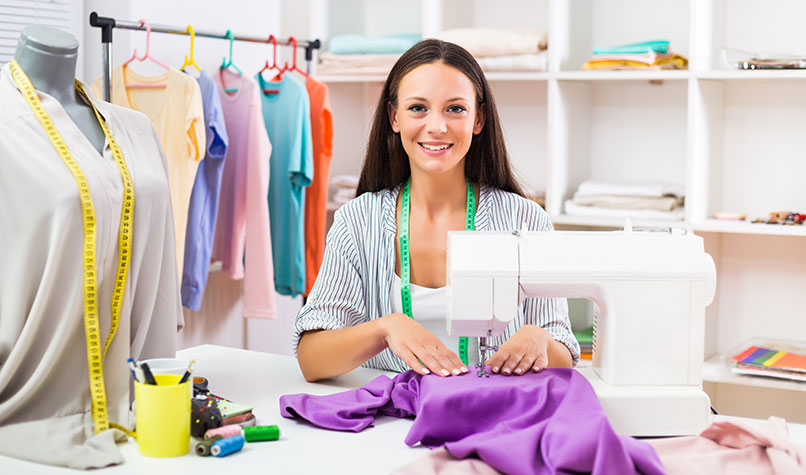 A woman dressmaker sat behind a sewing machine with colourful clothes behind her.