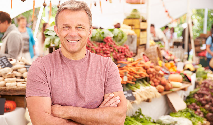 A greengrocer smiling with his arms crossed stood in front of his fruit and vegetable stall