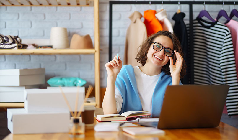 Woman in glasses sat a desk with a laptop with a rack of clothes behind her