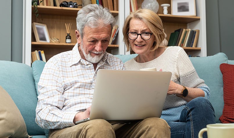 An older couple smiling sat on a sofa looking at a laptop.