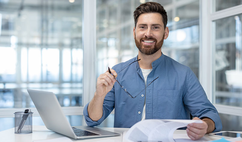 A bearded man sat at a desk with a laptop holding a pen