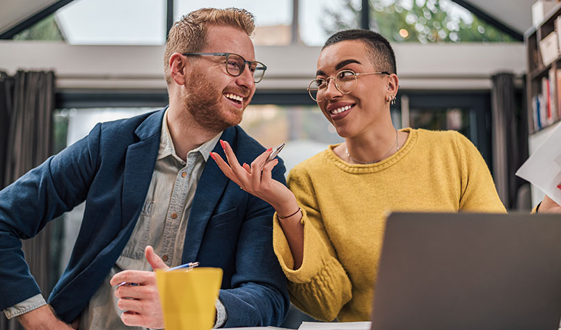 A man and woman smiling together sat at a desk with a laptop.