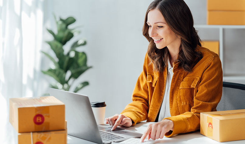 Woman wearing yellow sat at a desk with a laptop surrounded by boxes.