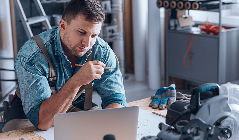 A carpenter in his workshop looking at a laptop on his workbench