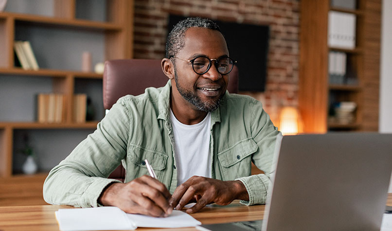 A man smiling and writing notes while watching his laptop screen