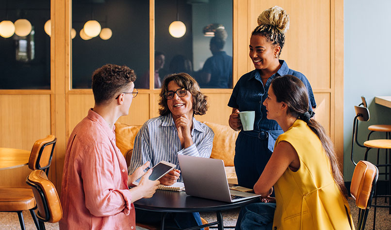 Four members of staff of various gender and ethnicity in an office canteen smiling while discussing something.