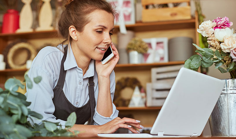 A female florist leaning on her sales counter looking at a laptop while she is on her mobile phone.