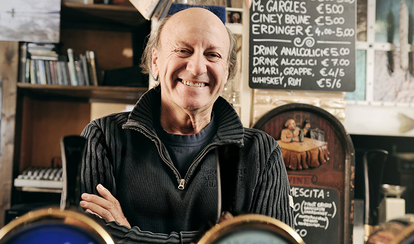 A middle aged male pub landlord smiling from behind a bar.
