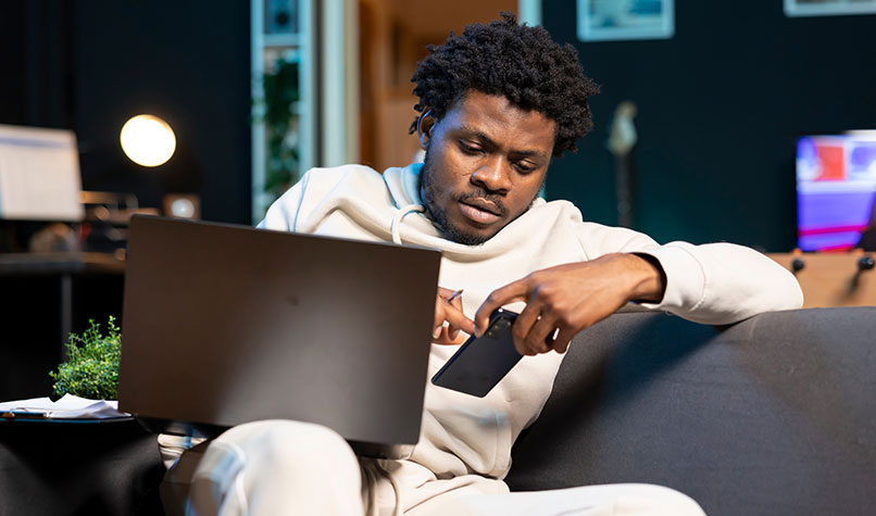 Man sat on his sofa looking at his phone with laptop on his lap.