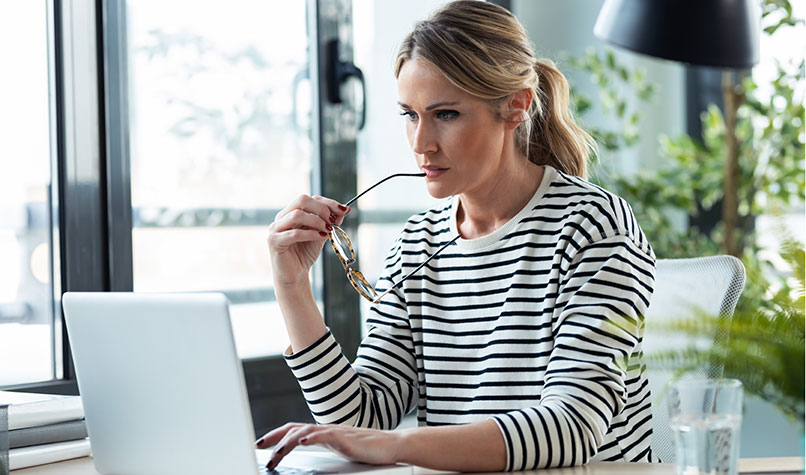 A blonde woman chewing her glasses while looking at her laptop computer.