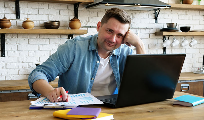 A man sat at kitchen table leading on his left hand with a laptop.
