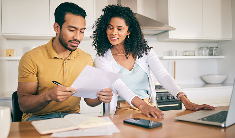 A man and a woman sat at a kitchen table looking at paper documents and a laptop.