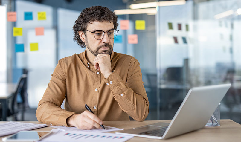 Man holding his chin sat in an office looking worriedly at his laptop