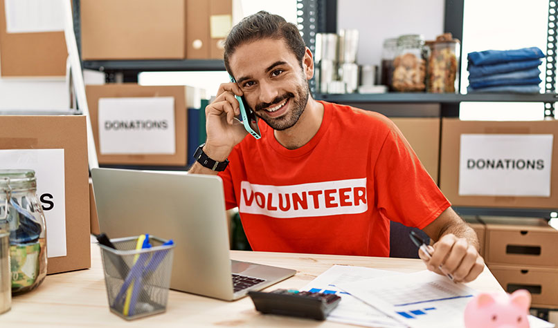 Man in a red T-shirt with 'Volunteer' written on it at a desk while on a telephone call.