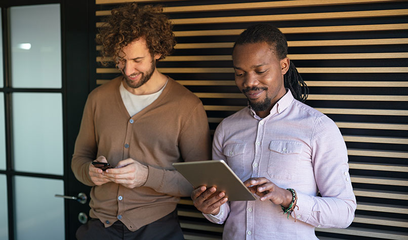 Two men leaning against a stripey wall smiling while looking at an iPad and mobile phone.