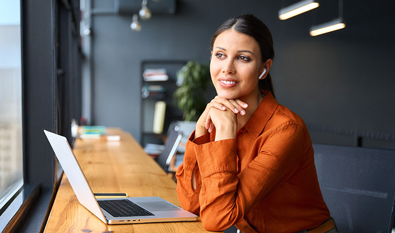 A woman looking out of a window with her laptop in front of her on her desk