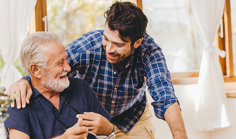 Young man hugging his dad who is sat at a dining table.