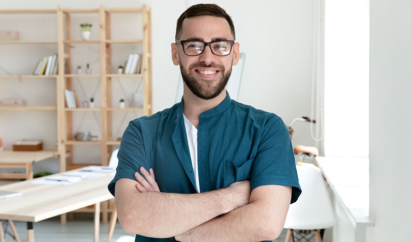A smiling man with beard and glasses with his arms crossed while standing in his home office.