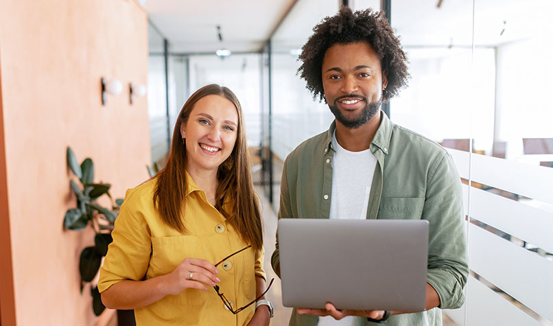 A woman wearing a yellow top and a man wearing a green top and holding a laptop smiling at the camera.