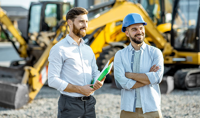 Two men standing in a construction site in front of a digger. The man on the right is wearing a blue hard hat and has his arms crossed.