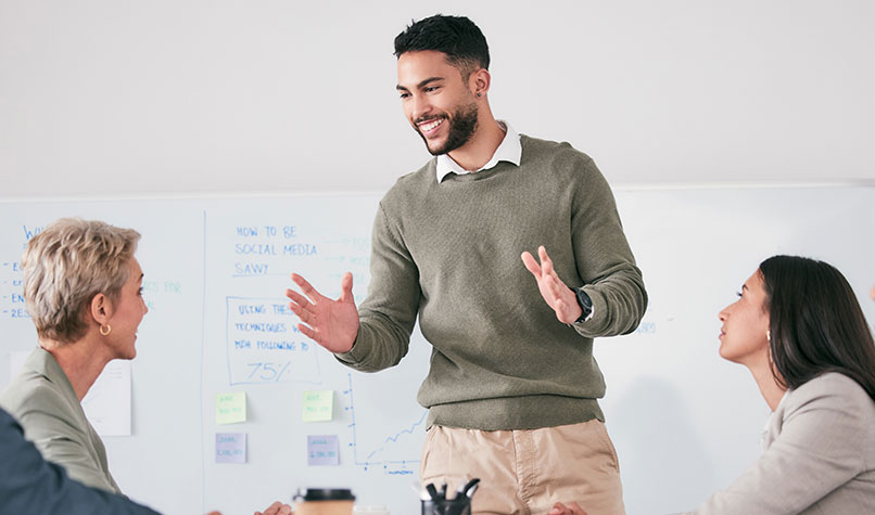 A bearded man standing up talking to a group or people sat around a meeting room table.