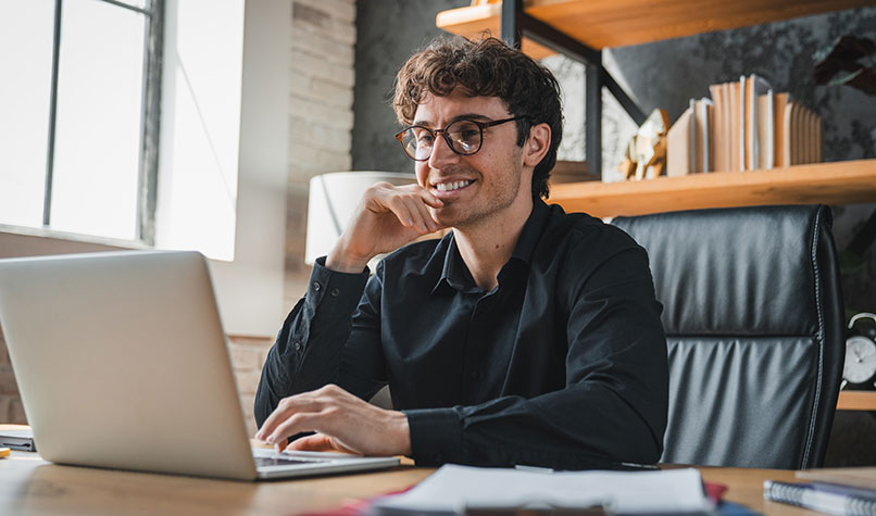A man wearing glasses sat at his desk and holding his chin while looking at his laptop
